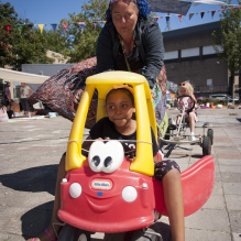 Girl in a small toy car being pushed by a woman with blue dreadlocks in Moerwijk, Den Haag